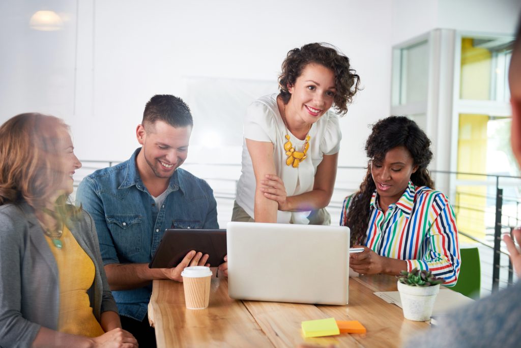 Four people sit around a table with a laptop, tablet, and coffee cup, collaborating and smiling in a modern office setting.