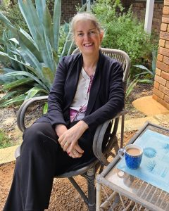 A woman sits on a chair outdoors next to a table with a blue mug of coffee, surrounded by plants and brick walls.