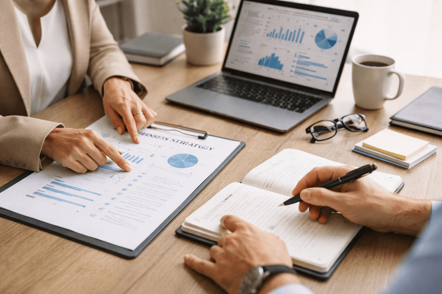 Two people discuss business strategy at a desk with documents, an open notebook, and a laptop displaying graphs and charts.