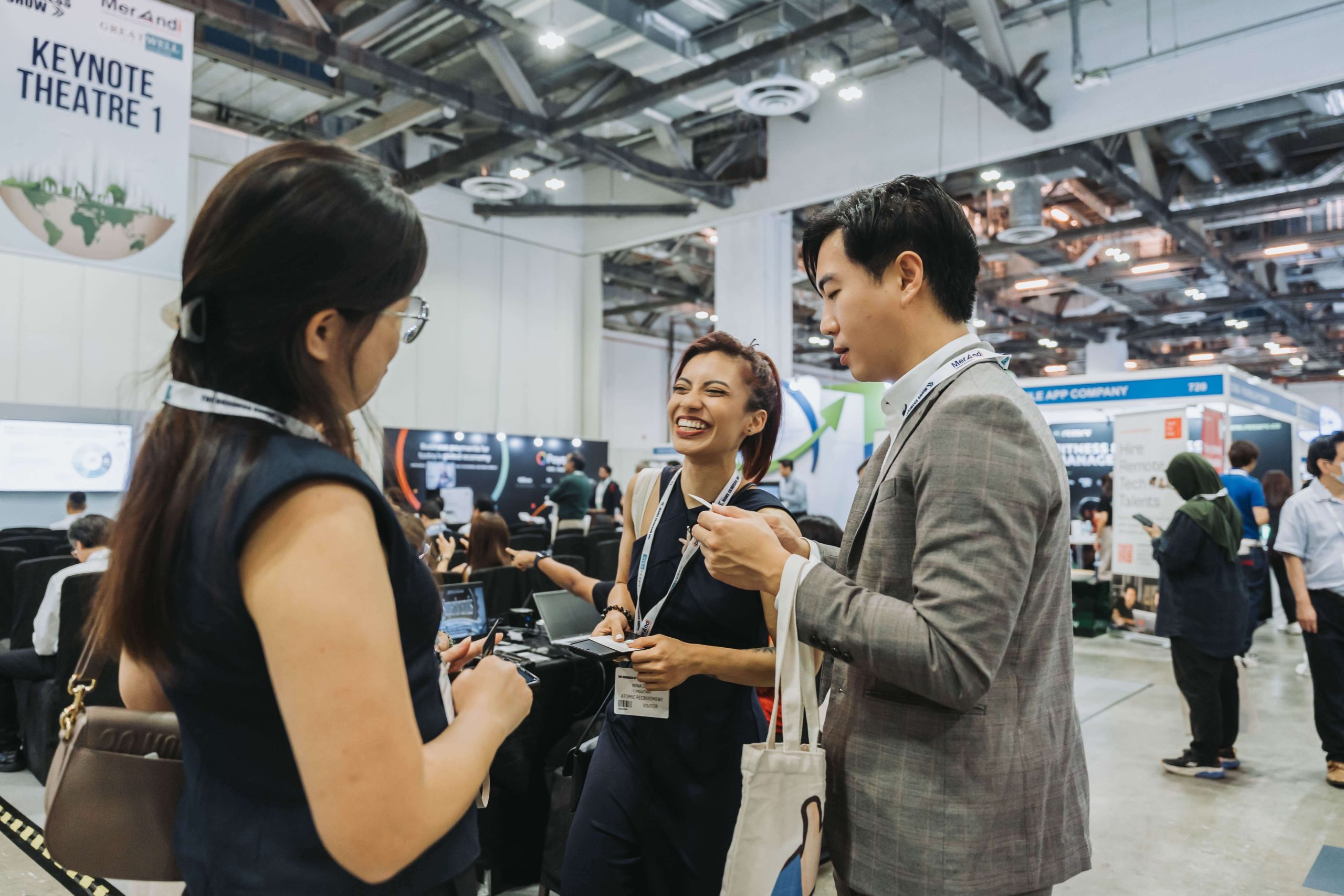 Three people wearing conference badges converse inside a busy exhibition hall, with booths and other attendees visible in the background.