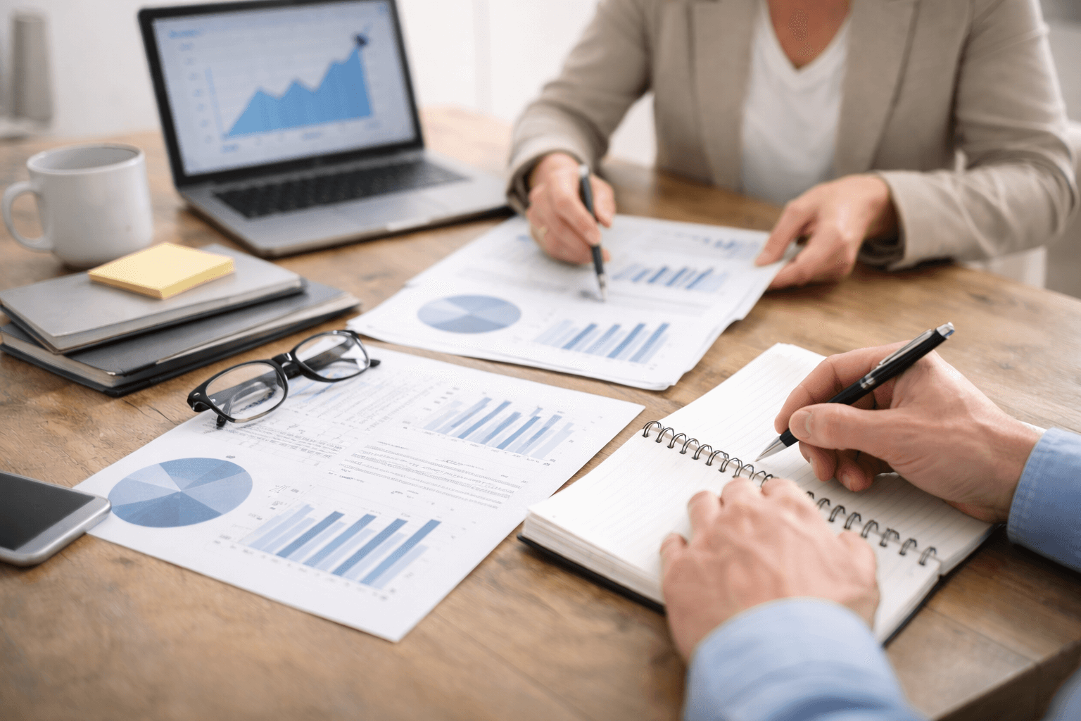 Two people review financial graphs and take notes at a desk with documents, a laptop showing a bar chart, glasses, and pens.