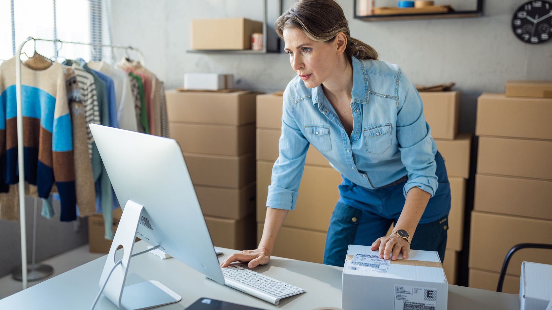 A woman stands at a desk, working on a computer while holding a package, with clothing and boxes visible in the background.
