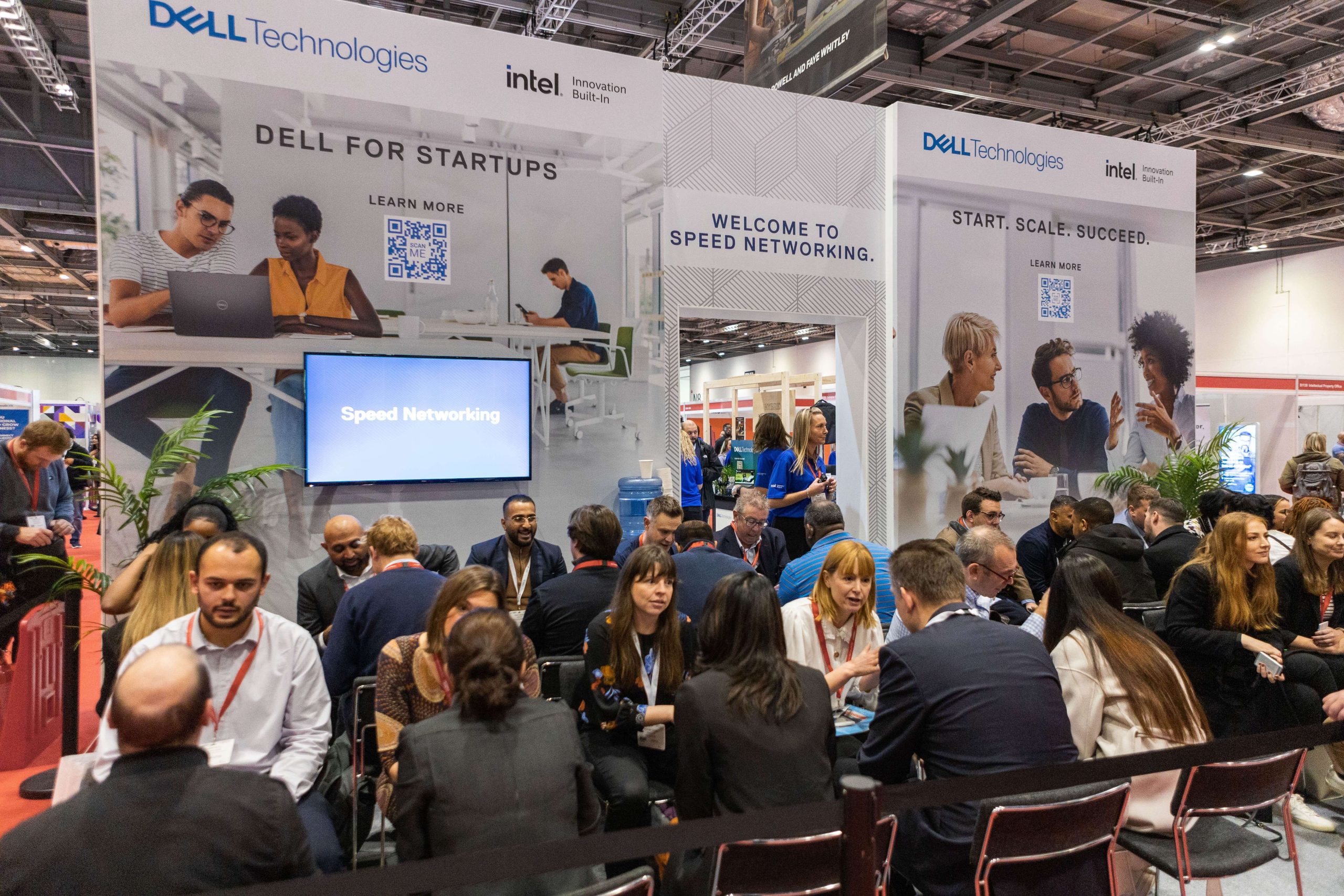People networking and conversing at a Dell Technologies booth during a business event, with banners promoting startup support and speed networking.