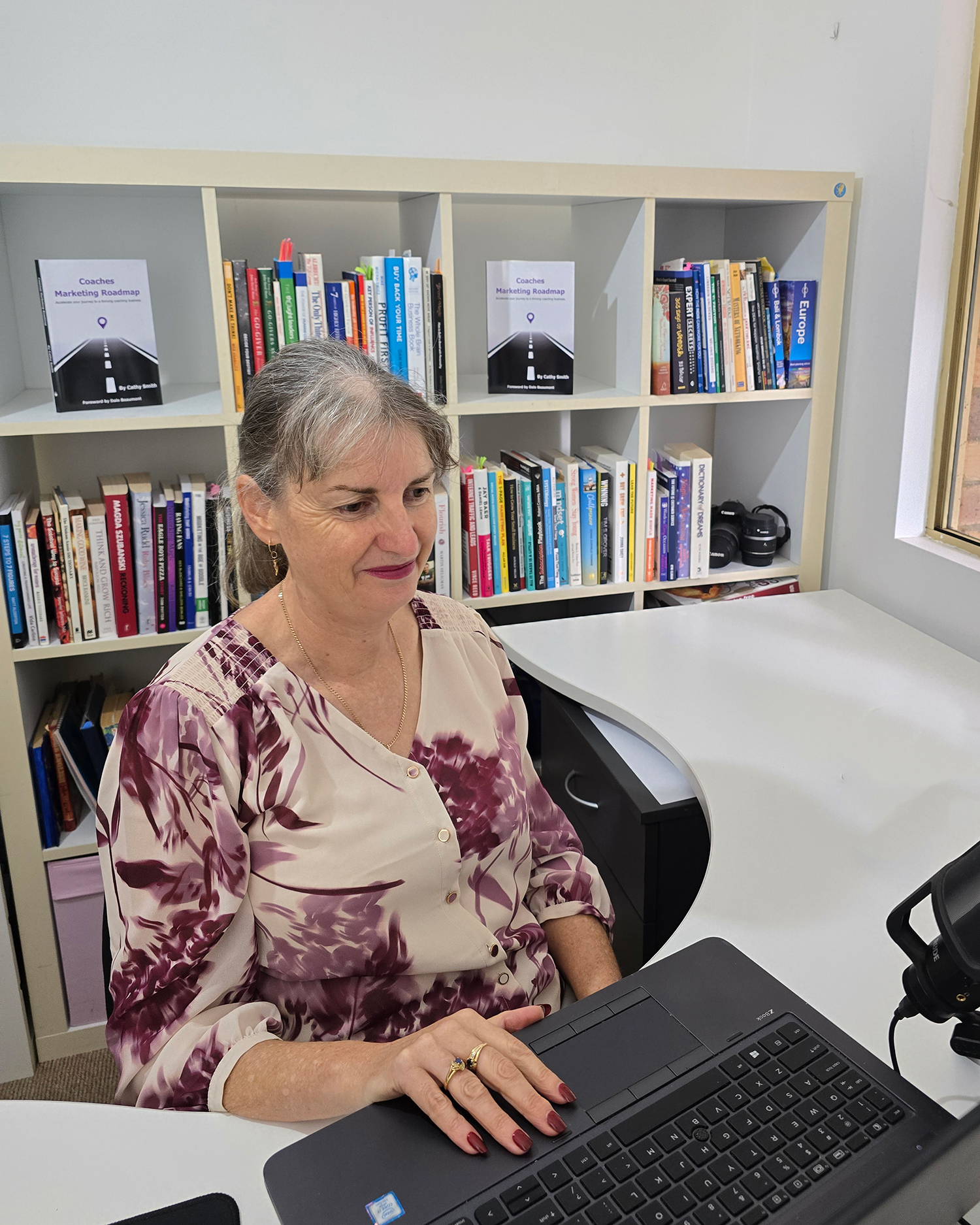 A woman sits at a desk using a laptop in an office with bookshelves filled with books and two copies of the same book displayed upright.