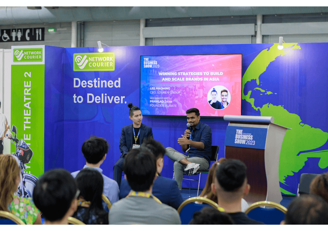 Two speakers sit on stage discussing business strategies at The Business Show 2023, with an audience seated in front and a large branded backdrop behind them.