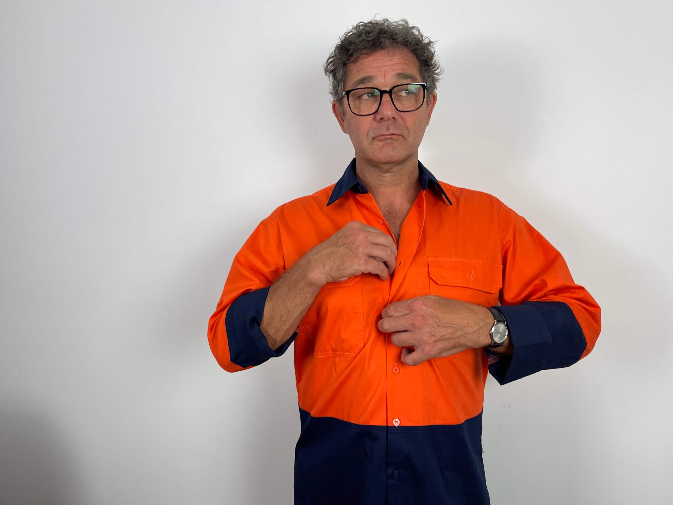 A man wearing glasses and an orange and navy work shirt stands against a white background, buttoning his shirt.