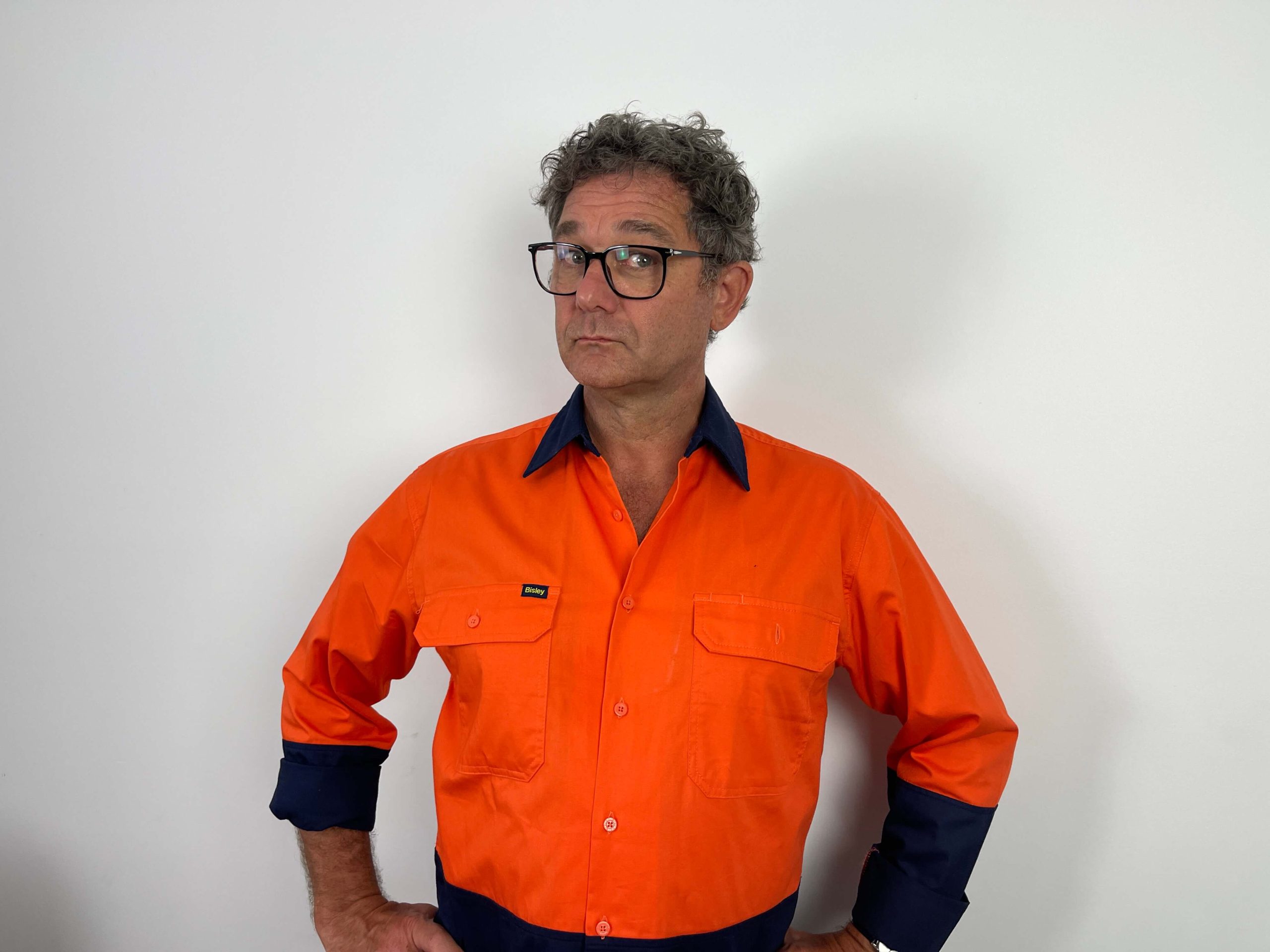 A man wearing glasses and an orange work shirt with dark blue cuffs stands against a plain white background with his hands on his hips.