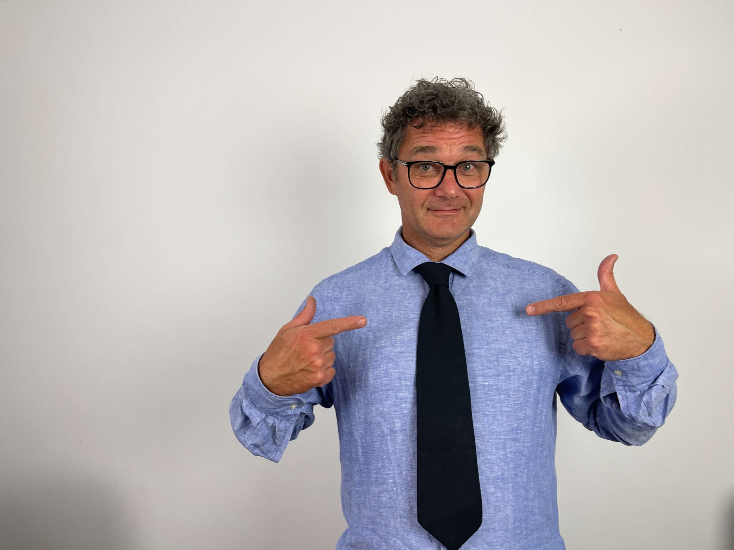 Man wearing glasses and a blue shirt with a black tie stands against a plain white background, pointing at himself with both hands.