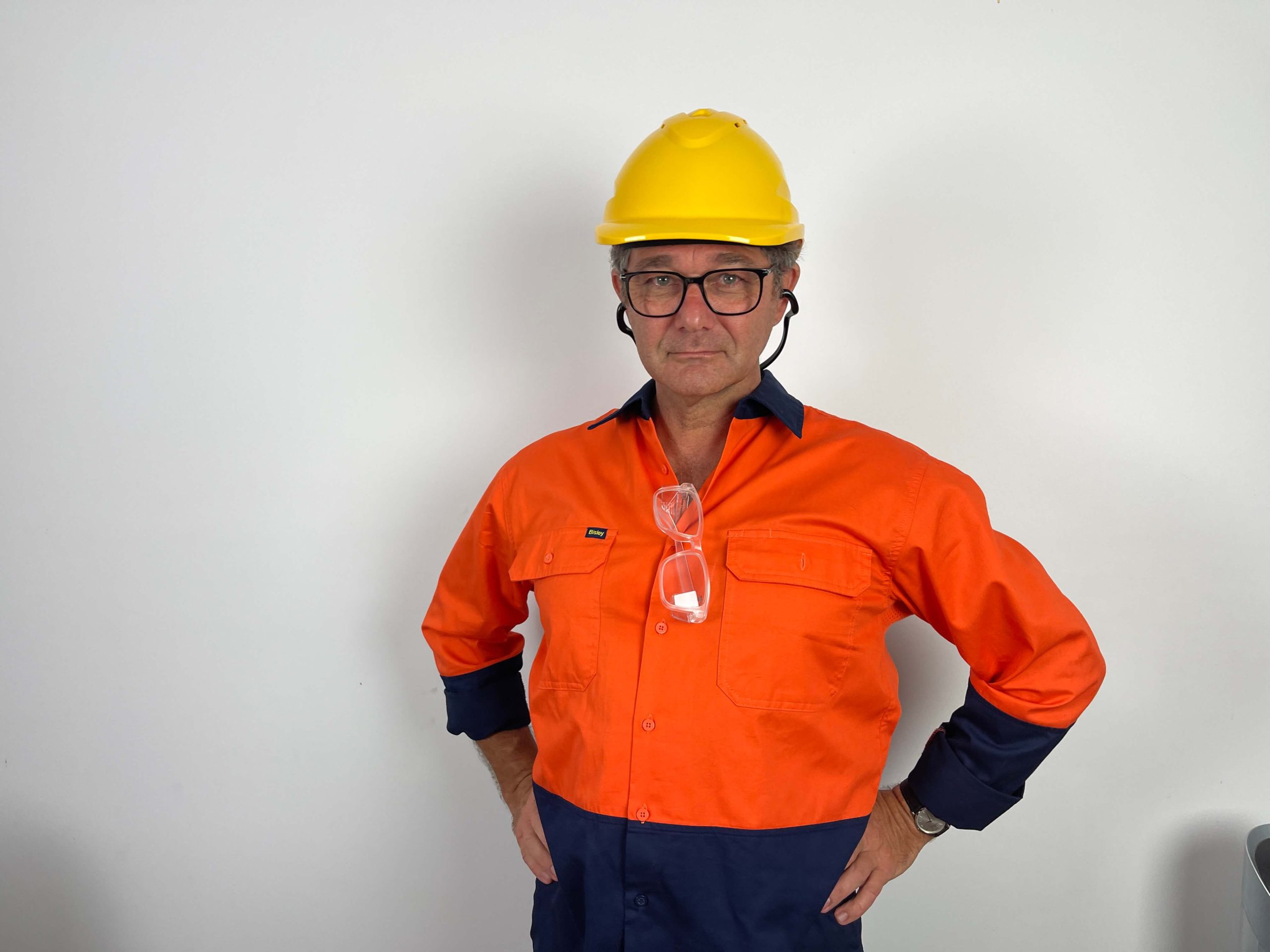 A man wearing a yellow hard hat, glasses, and an orange and navy work shirt stands against a plain white wall with his hands on his hips.