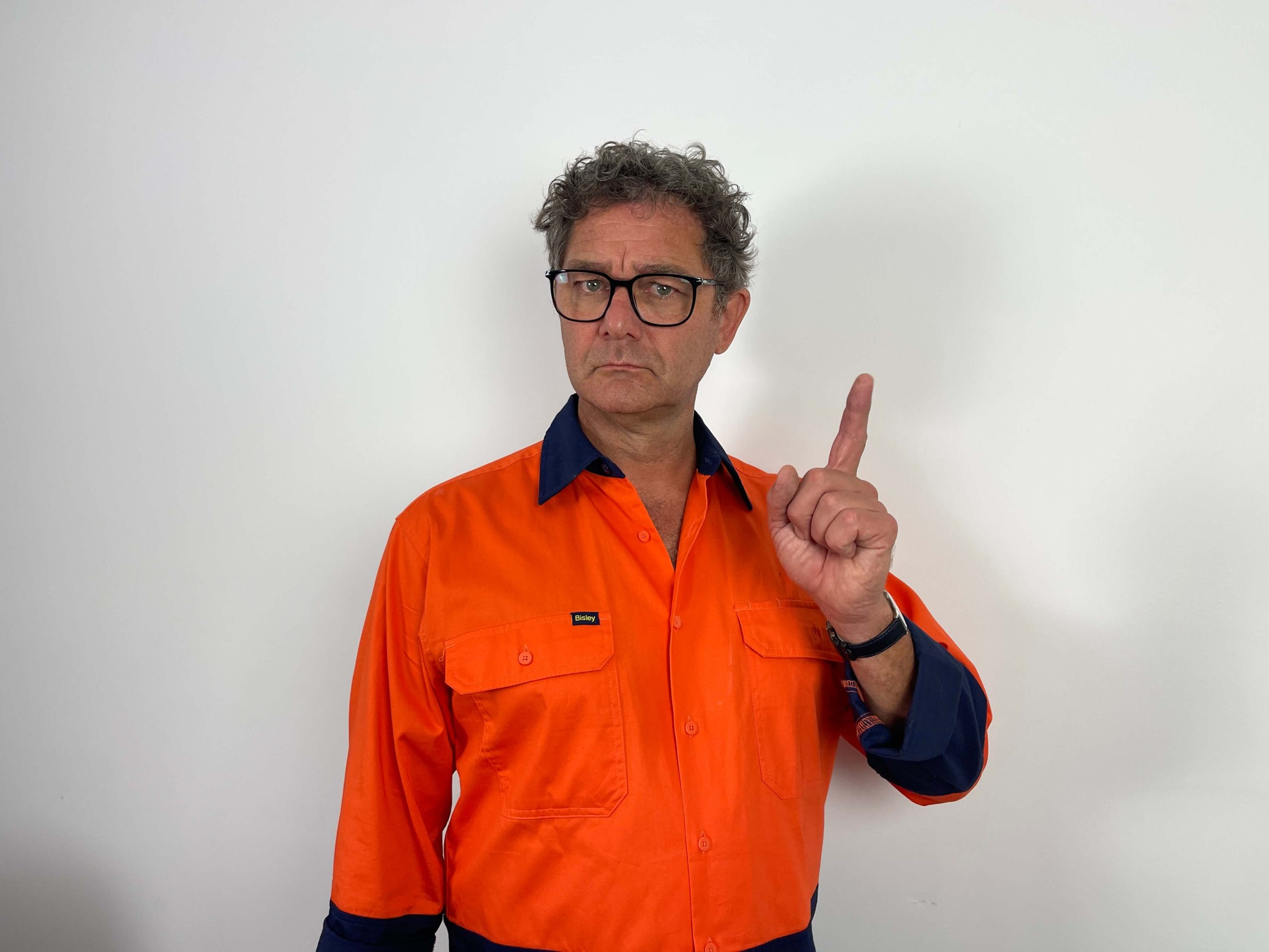 Man in an orange work shirt and glasses stands against a white background, holding up his right index finger in a gesture.