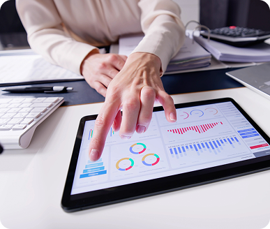 A person points at a tablet displaying various colorful graphs and charts, with a keyboard and papers on the desk nearby.