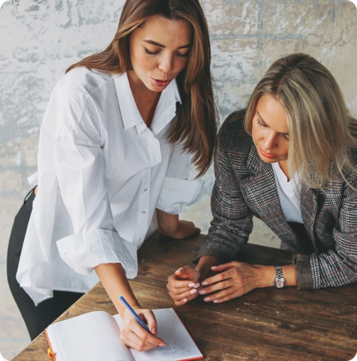 Two women at a wooden table look at an open notebook; one is writing while the other observes, both engaged in discussion.