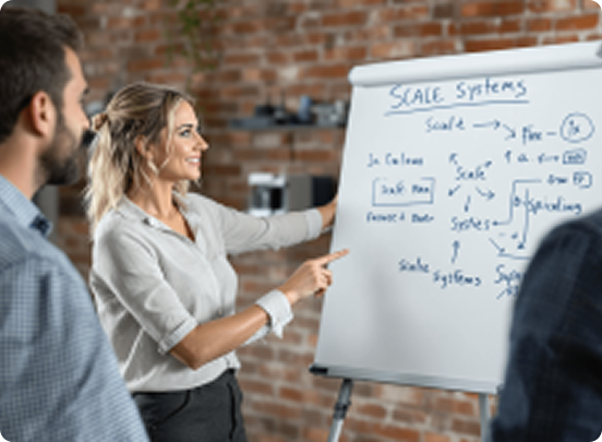 A woman points to a flowchart on a flip chart labeled "Scale Systems" during a meeting with two men in a brick-walled room.