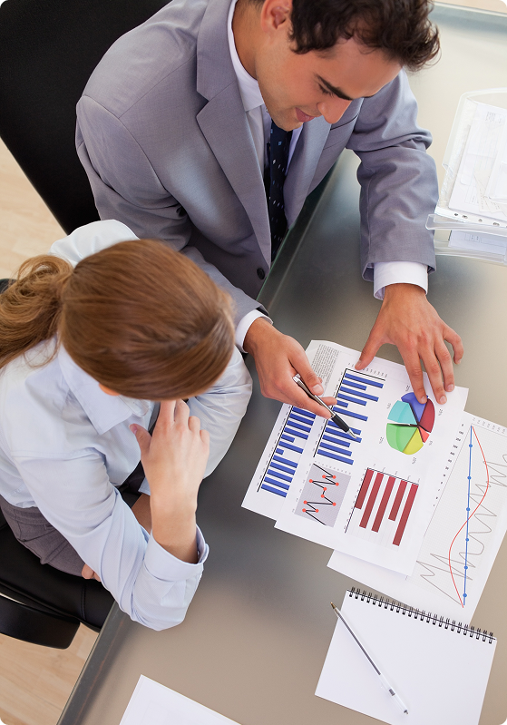 Two people in business attire discuss graphs and charts at a glass table, analyzing data on printed sheets.