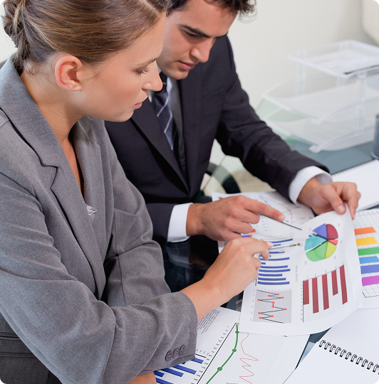 Two business professionals analyzing graphs and charts at a desk, using a pen to point at data on the documents.